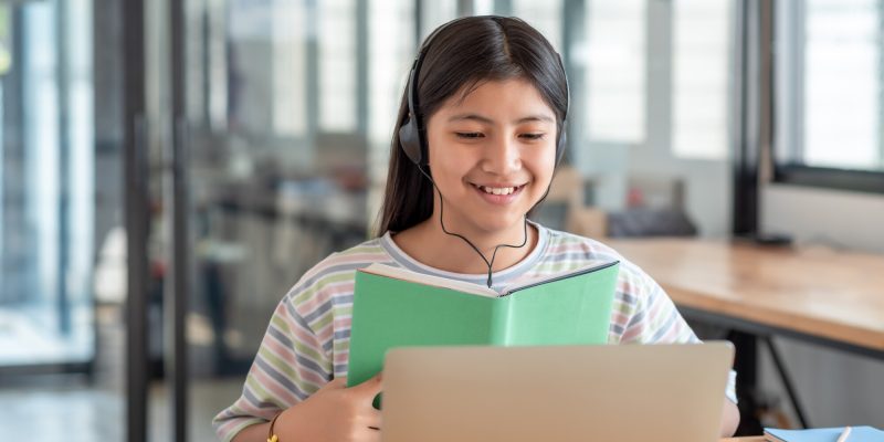Image of a happy Asian girl wearing headphones holding a textbook online learning using a laptop in