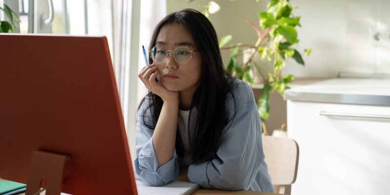 Young focused Asian girl student watching webinar video course on computer during online learning